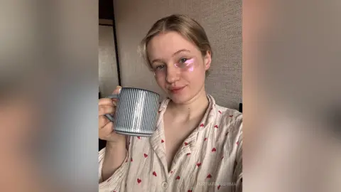 Blonde woman in white patterned pajamas holding a striped mug in a cozy, relaxed indoor setting.