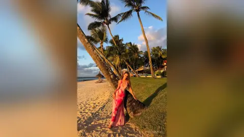 Woman in a long pink floral dress standing on a sunny tropical beach near palm trees at sunset.