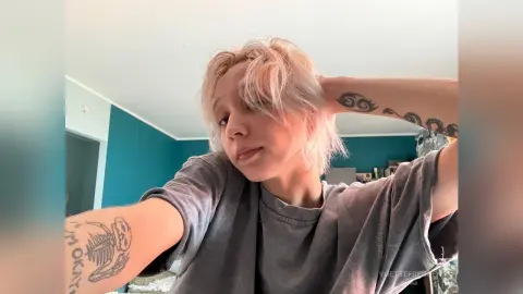 Young woman with blonde hair and arm tattoos wearing a grey t-shirt in a teal bedroom.