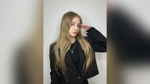 Blonde woman in a black blazer and necklace saluting against a white background with a confident mood.