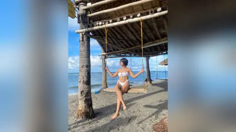 Young woman in a white bikini relaxing on a wooden swing under a thatched beach hut by the ocean.