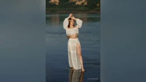 StelaBrown stands in shallow water wearing a white off-the-shoulder crop top and a long slit skirt with her hands behind her head.