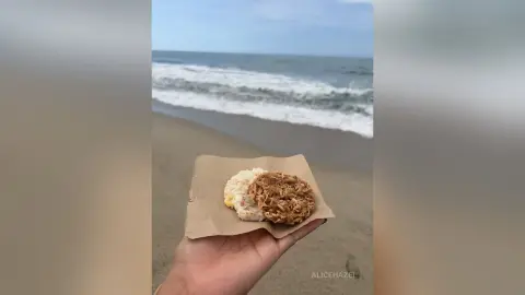Hand holding a snack of rice and fried fritters on a paper plate at a sunny beach by the ocean.