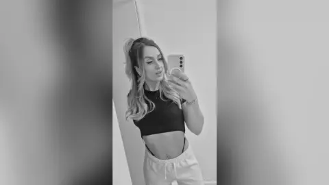 Young woman in a black crop top and white sweatpants taking a moody black and white mirror selfie.