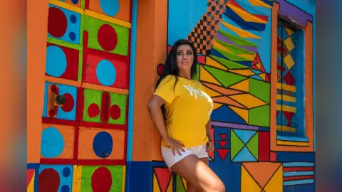 MissyJolie poses against a bright, multi-colored geometric wall wearing a yellow t-shirt and white shorts.