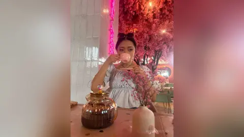 Young woman in a white blouse sipping tea in a dreamy pink cherry blossom cafe setting.