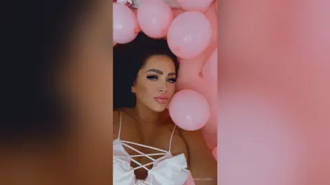 Glamorous woman in a white satin dress posing amidst soft pink balloons for a dreamy birthday celebration.