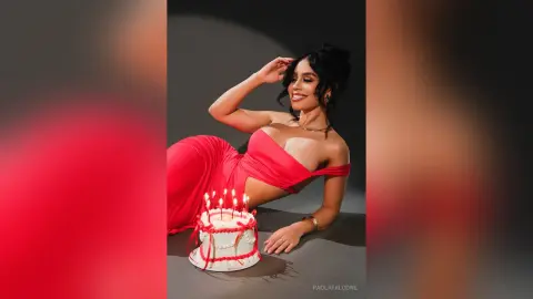 Smiling woman in a red dress lying next to a white birthday cake in a moody studio setting.