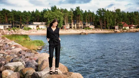 Elegant woman in a black jumpsuit standing on rocky shores by a serene lake surrounded by pine trees.