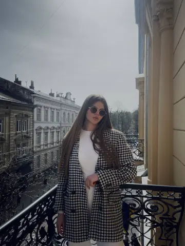 Stylish woman in a houndstooth coat and sunglasses posing on a balcony overlooking classic European architecture.