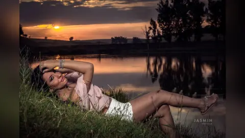 MissyJolie reclines on a grassy bank by a lake at sunset, wearing a light pink ruffled top and white bottoms.