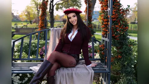 Smiling woman in a burgundy cardigan and beret sitting on a park bench amidst autumn flowers.