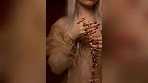 EllieFrosty poses with her hands near her face, wearing gold jewelry and red nail polish against a dark background.