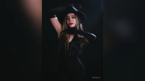 JessieLiu poses in a black outfit and black gloves against a dark background, wearing a wide-brimmed black hat.