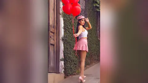 Stylish woman in a pink skirt and white crop top holding red heart balloons against a rustic wall.