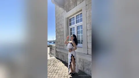Elegant woman in a white sleeveless maxi dress posing by a stone building under a clear blue sky.