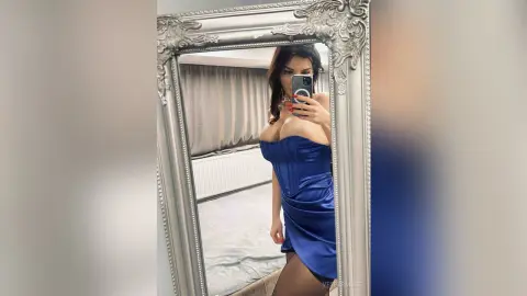 VesperMuse poses in a mirror wearing a tight, strapless blue satin dress that shows off her cleavage and curves.