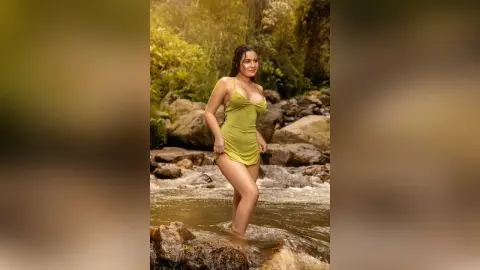 Young woman in a lime green slip dress standing in a sunlit forest river surrounded by rocks.