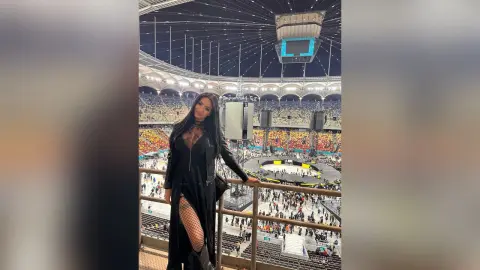 Woman in a long black dress and fishnet tights standing on a balcony at a crowded music concert stadium.