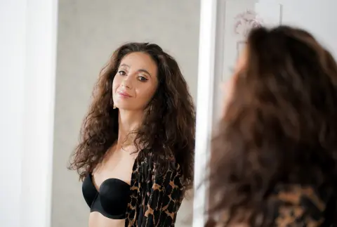 Smiling woman with curly brown hair wearing a black bra and leopard print robe in a bright room.