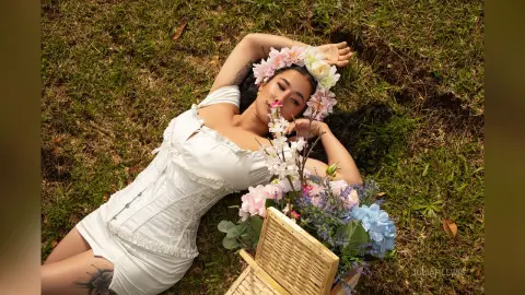 JuliahLewis lies on green grass wearing a white lace corset and a floral headpiece, surrounded by pink and blue flowers.