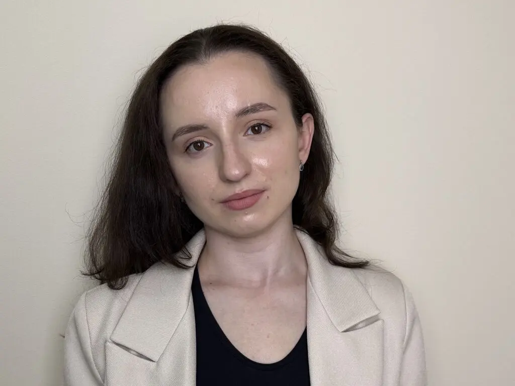 Young woman with brown hair wearing a beige blazer and black top against a neutral background.