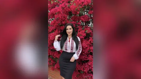 JasminNise poses with long black hair, wearing a white long-sleeve blouse with red embroidery and a black skirt against a wall of bright red flowers.