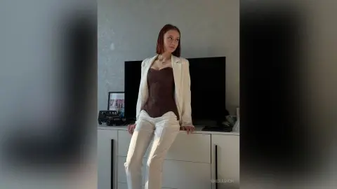 SheraRich poses in a white blazer and matching white pants, wearing a dark brown corset while leaning against a white dresser in front of a TV.