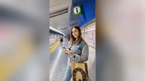 JessicaAidennn — Young woman in a denim jacket holding a phone and shopping bag in a bright subway station.