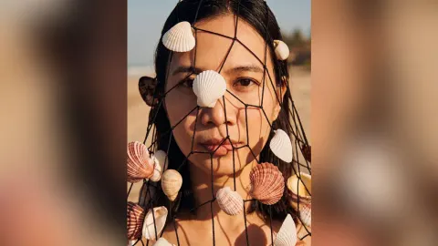 MilaSwan — MilaSwan looks at the camera through a black net decorated with white seashells on a sunny beach.