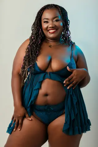 Smiling Black woman in teal lace lingerie and flowing robe posing against a neutral studio background.