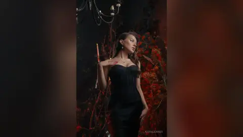 Elegant woman in a black corset dress posing in a moody, dark floral garden setting with candlelight.