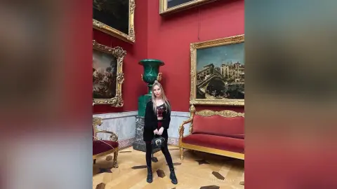 Young woman in a black outfit standing in a classic art gallery surrounded by gold framed paintings.