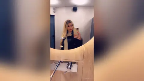Blonde woman in a black leather jacket taking a mirror selfie in a modern bathroom setting.