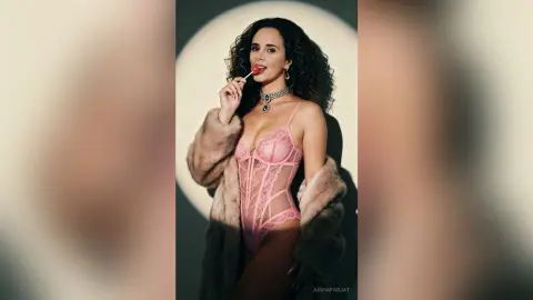 Glamorous woman in pink lace bodysuit and fur coat eating a lollipop against a studio backdrop.