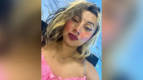 Young woman with glamorous makeup and blonde hair wearing a pink lace top in a bright setting.