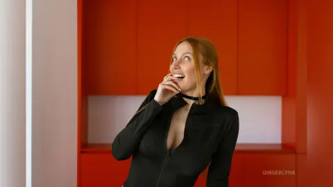 Smiling redhead woman in a black long-sleeve top posing against a vibrant red cabinet background.