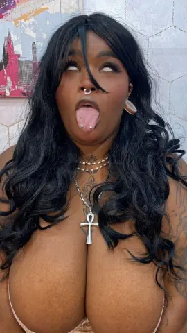 Curvy woman with long wavy black hair and ankh necklace posing playfully in a bright room.