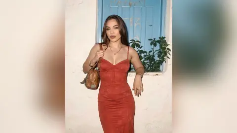 Stylish woman in a red midi dress and gold jewelry posing against a rustic white wall and blue door.