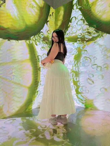 Smiling woman in a white maxi skirt and black tank top standing against a vibrant lime citrus mural background.
