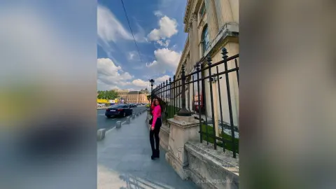 Stylish woman in a pink top and black trousers walking on a city sidewalk near historic architecture under a blue sky.