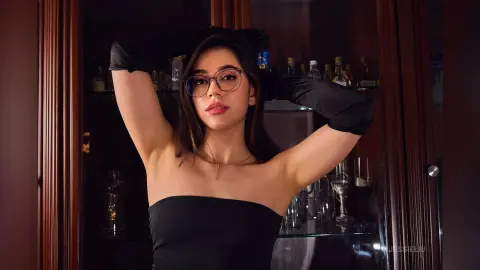 JessieLiu poses with her hands behind her head, wearing a black strapless top and black gloves against a dark wooden bar background.