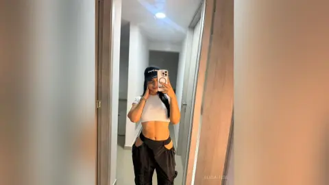 Young woman in white crop top and black cargo pants taking a mirror selfie in a modern hallway.