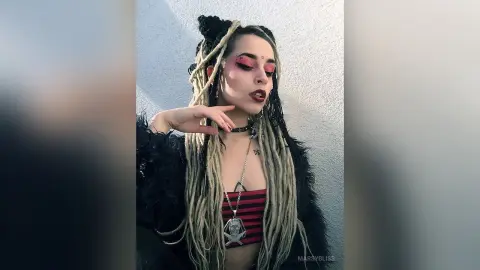 Woman with long blonde dreadlocks wearing a black faux fur coat and silver skull necklace against a grey wall.
