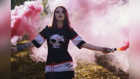 Gothic woman in black graphic t-shirt holding pink smoke bomb in a forest setting.