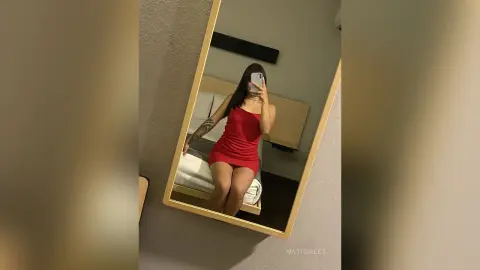 NatiSweet poses in a short red dress while looking into a mirror, showing her long dark hair and arm tattoo.