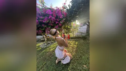 Blonde woman in a white summer outfit relaxing on a sunny garden lawn near blooming pink bougainvillea.