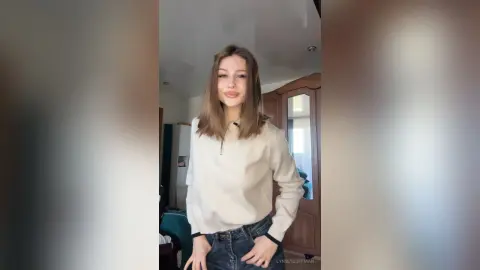 Smiling young woman in a white long-sleeve shirt and blue jeans posing in a bright living room.