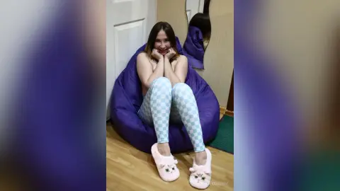 Smiling young woman in checkered leggings and pink fuzzy slippers relaxing on a purple bean bag chair.