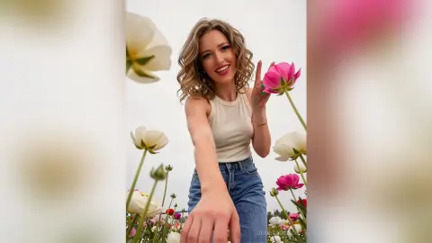 Smiling young woman in a white tank top and jeans posing amidst large white and pink flowers in a bright field.